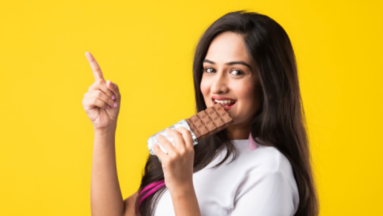 Portrait of a happy young Indian asian young woman biting chocolate bar isolated over pink or yellow background