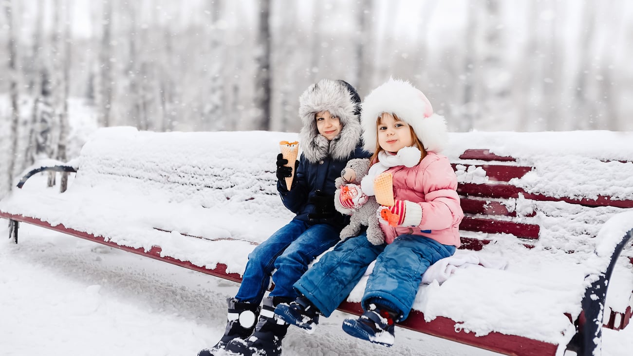 Two young children eat ice cream dressed in winter gear, sitting on a park bench full of snow.