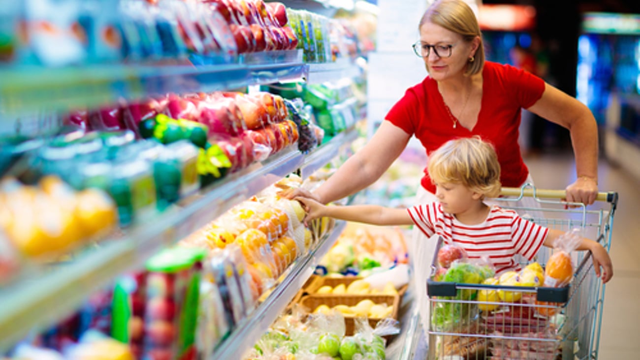 Adult woman pushes a grocery store basket with a child in it, both reach for produce from a grocery store fridge.