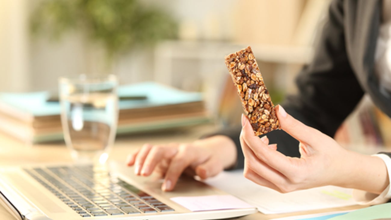 Close up of entrepreneur woman hands holding cereal snack bar working on laptop at home office