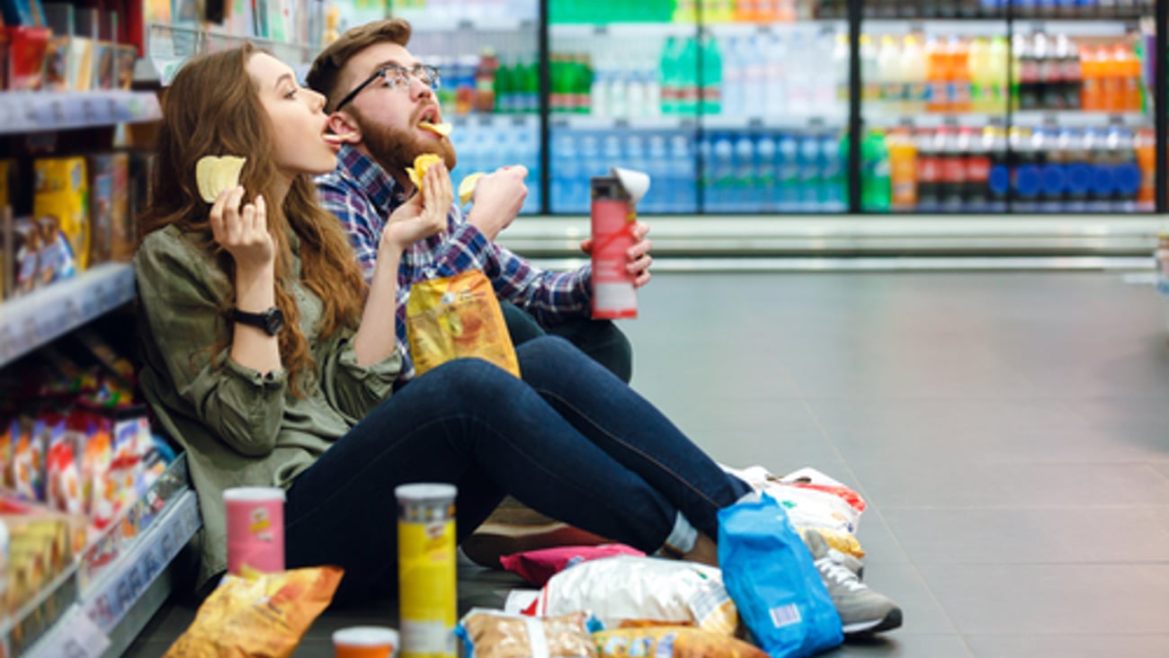 Portrait of a young funny hungry couple sitting on the supermarket floor and eating junk food