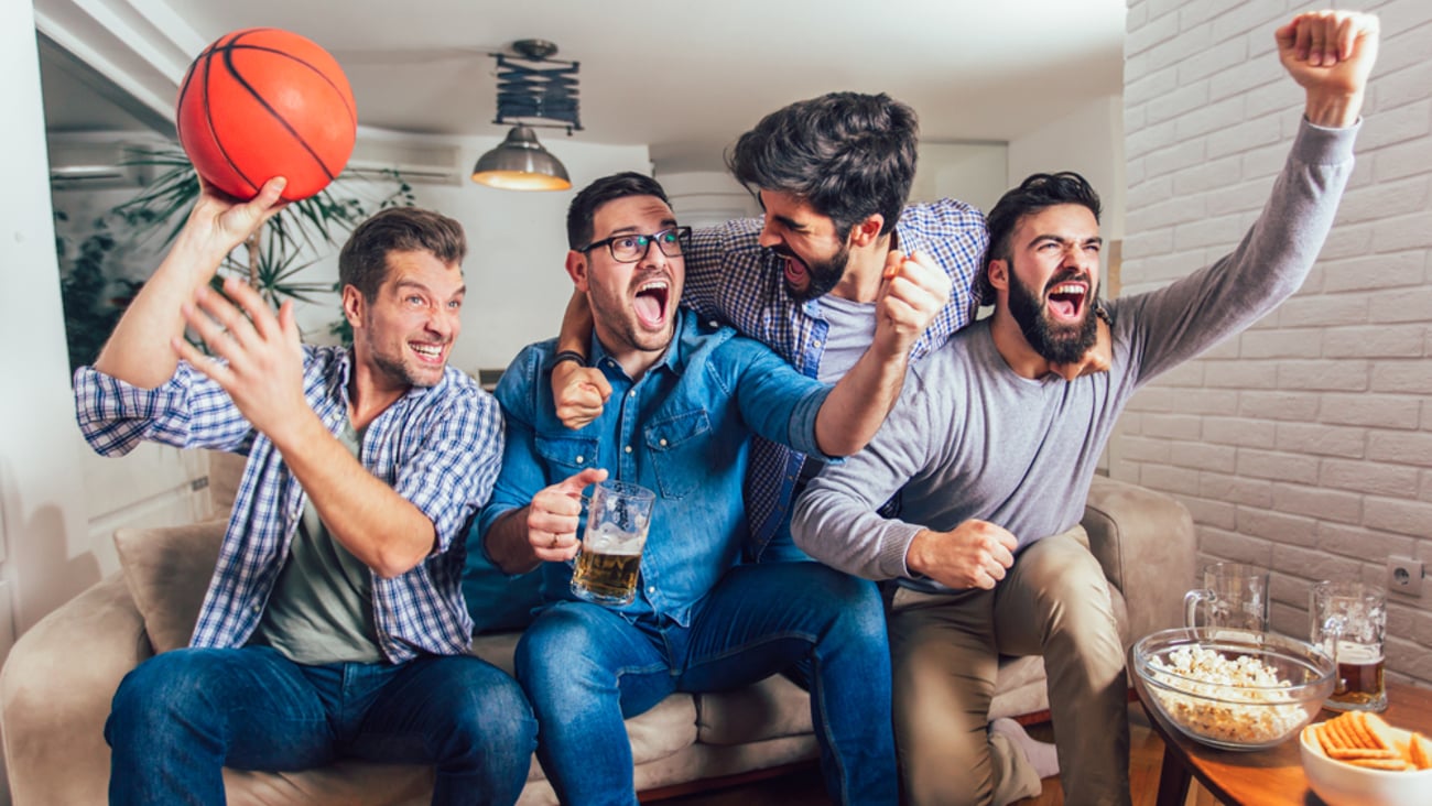 A group of cheering friends on a couch with popcorn, chips and beer in front of them. One is holding a basketball.