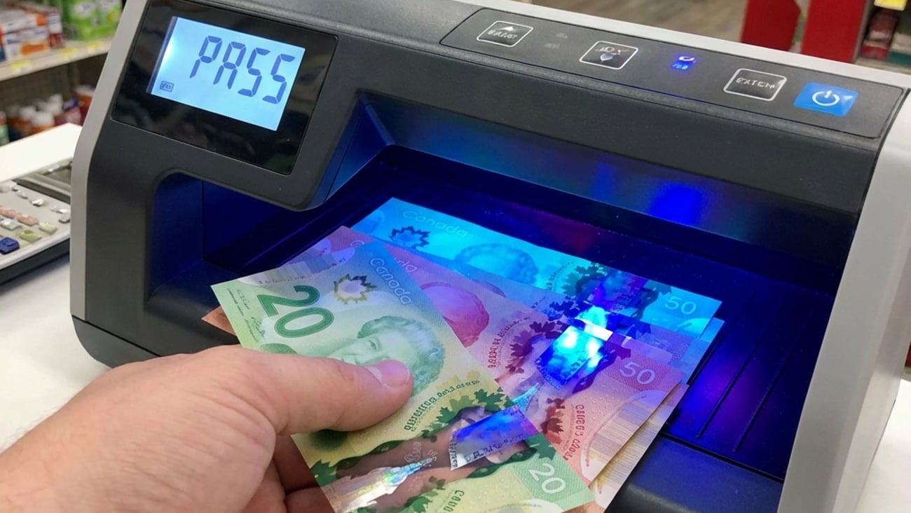 A clerk is sliding Canadian bills into a counterfit detection machine that reads "pass".