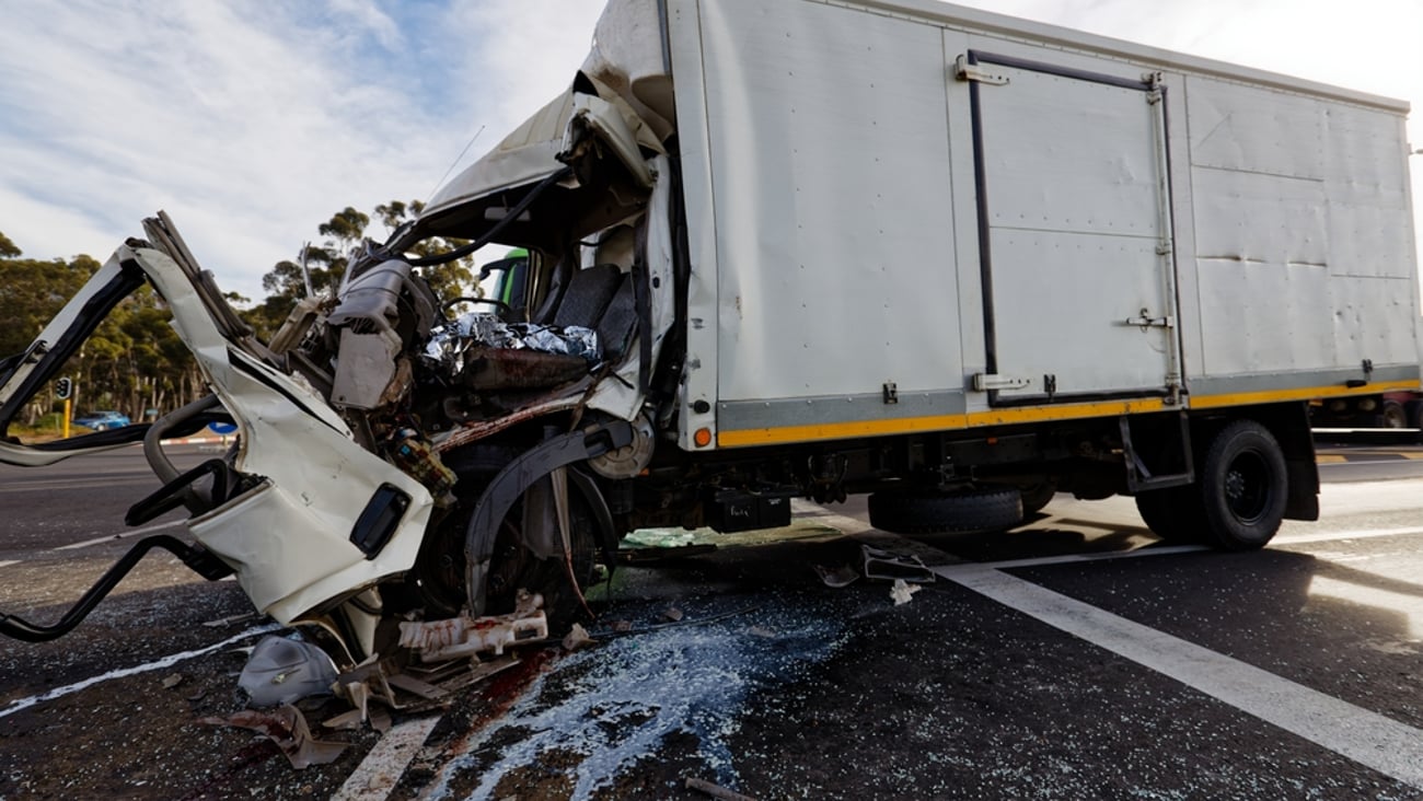 An overtuned truck with its front cab badly damaged in impact; liquid is leaking out from underneath the mangled truck.