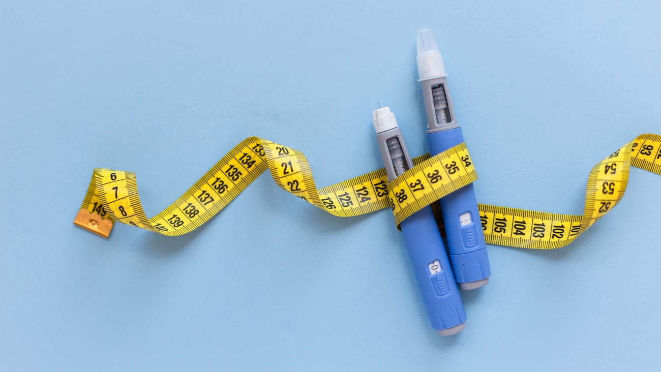 Two injectors dosing pens for subcutaneous injection of antidiabetic medication or anti-obesity medication hovering over a blue background. Yellow measuring tape around the injectors. 