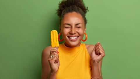 Happy dark skinned millennial girl eats yellow ice cream of mango flavor, clenches fist with anticipation of something awesome happen, has toothy smile, wears bright summer clothes, isolated on green