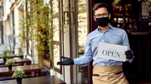 Happy waiter with protective face mask holding open sign while standing at cafe doorway.