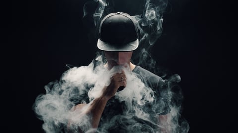 Man smoking a vape, surrounded by smoke, on black background wearing black baseball cap. 