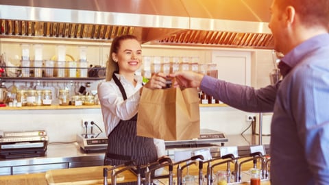 Cheerful waitress wearing apron serving customer at counter in restaurant - Small business and service concept with young woman owner offering recycled paper bag with take away food to online client