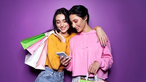 Two happy women friends shoppers holding shopping bags using mobile apps for online shopping standing on purple background. Retail ecommerce fashion sale offers, mall discounts in applications concept