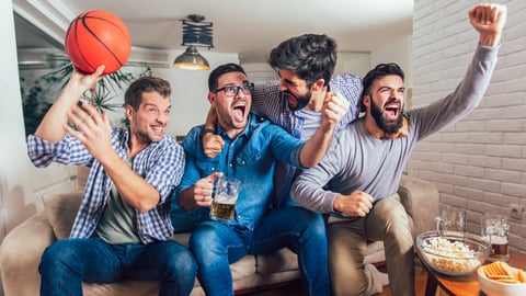 A group of cheering friends on a couch with popcorn, chips and beer in front of them. One is holding a basketball.