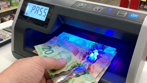 A clerk is sliding Canadian bills into a counterfit detection machine that reads "pass".
