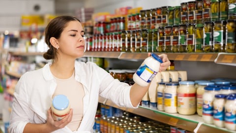 Young dark-haired woman at store shelves holding mayonnaise jar, reading label