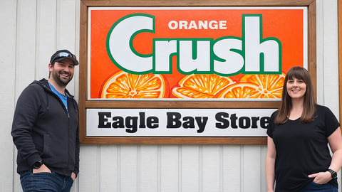 Eagle Bay Store Owners Brittany and Dave Huston in front of their sign that features an ad for Orange Crush