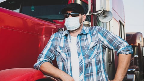 Semi truck professional driver on the job in casual clothing wears safety medical face mask. Confident looking trucker stands next to red big rig wearing protection sunglasses and surgical mask.