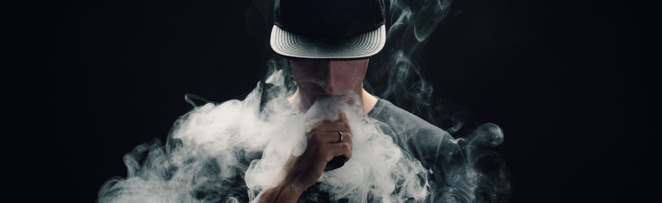 Man smoking a vape, surrounded by smoke, on black background wearing black baseball cap. 