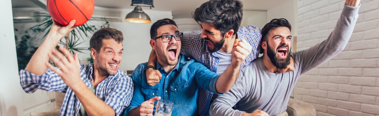 A group of cheering friends on a couch with popcorn, chips and beer in front of them. One is holding a basketball.
