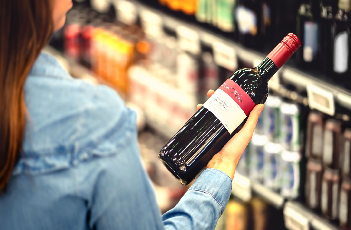 Woman reading the label of red wine bottle in liquor store or alcohol section of supermarket. Shelf full of alcoholic beverages. Female customer holding and choosing a bottle of merlot or sangiovese.