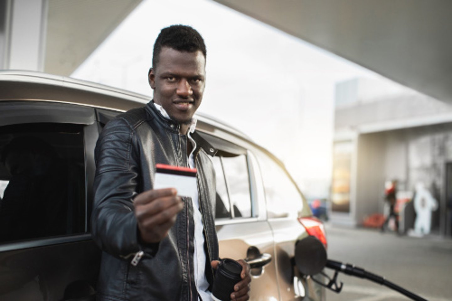 Cheerful young handsome black man smiling to camera and demonstrating his credit card before paying for gasoline on modern petrol station. Cashless payment concept