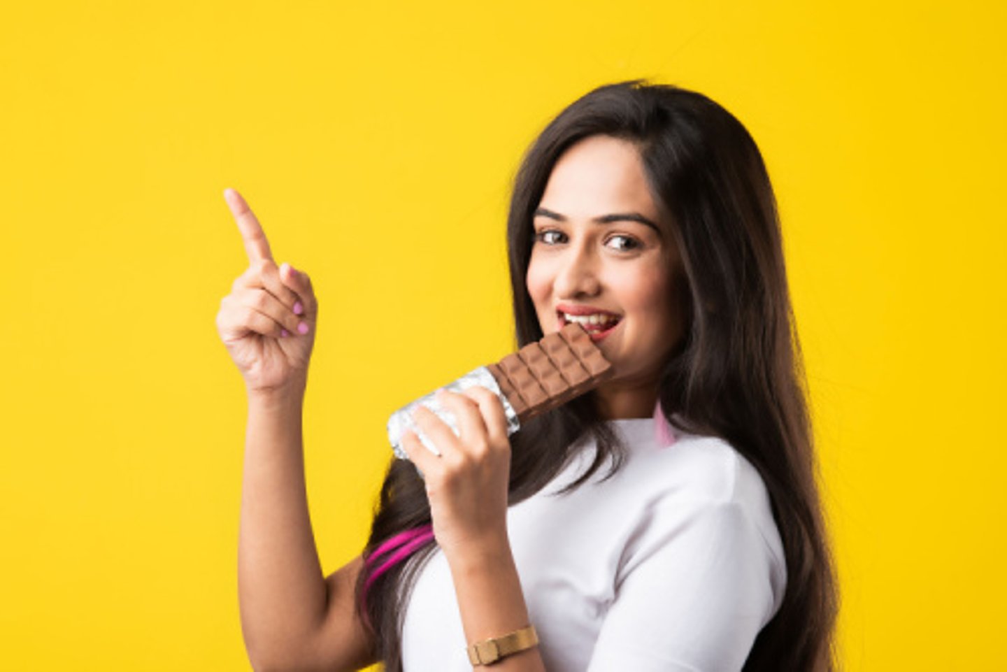 Portrait of a happy young Indian asian young woman biting chocolate bar isolated over pink or yellow background