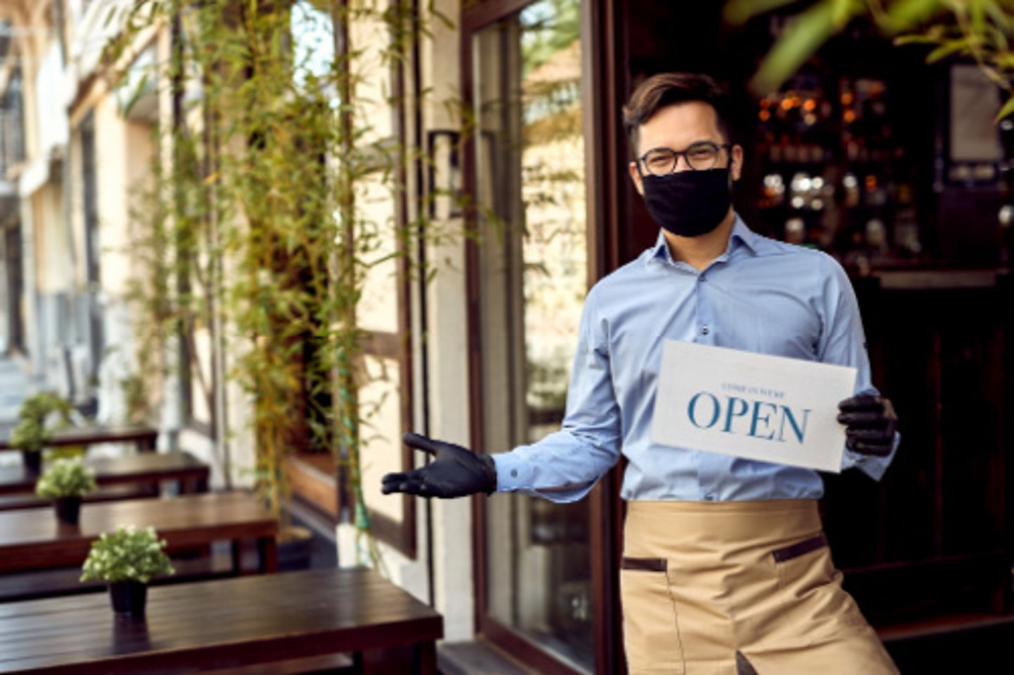 Happy waiter with protective face mask holding open sign while standing at cafe doorway.