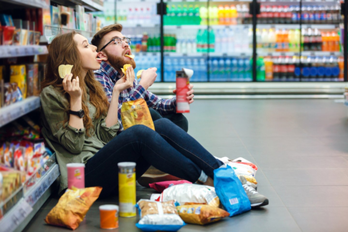 Portrait of a young funny hungry couple sitting on the supermarket floor and eating junk food