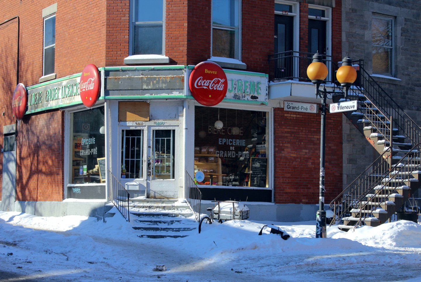 2019: A winter street on the Plateau Mont Royal with an old convenience store and soda commercials, a lamppost and an outside staircase in front of a red brick house