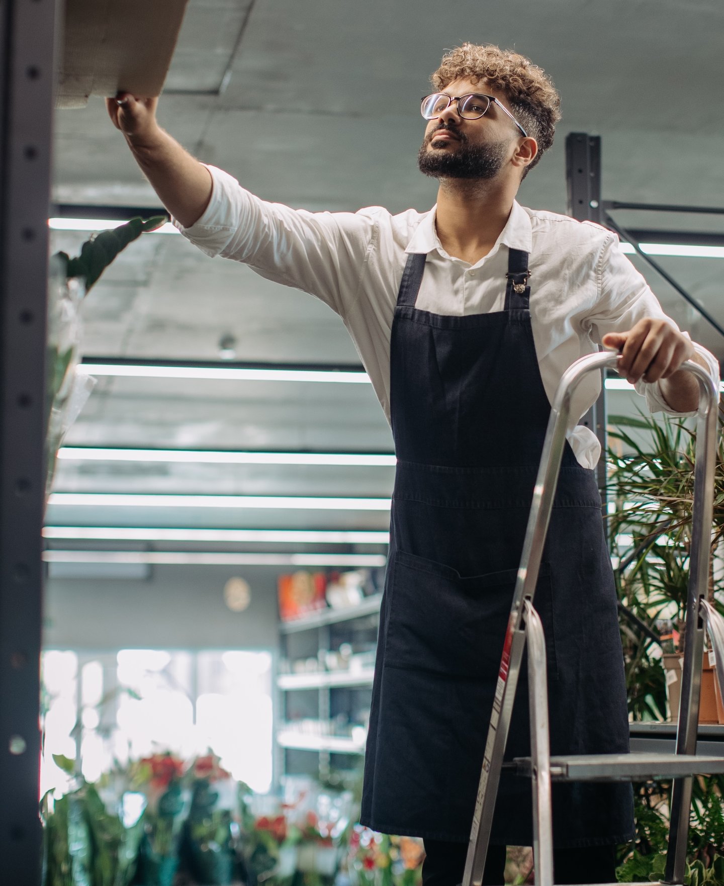 Male worker in apron in store or ladder reaching for top shelf