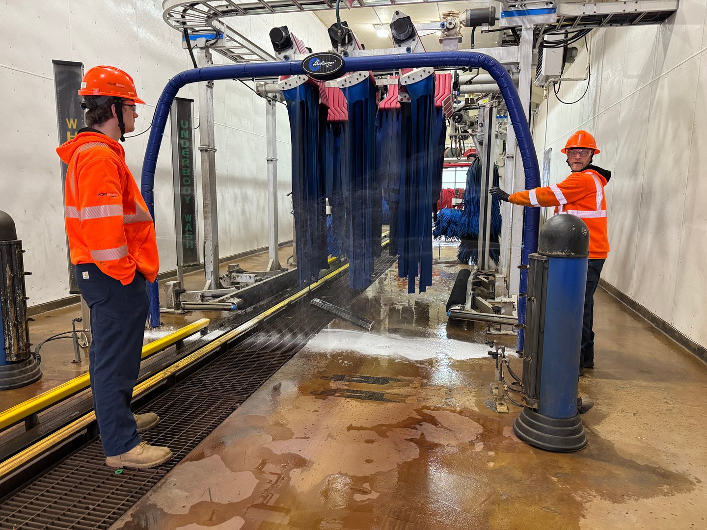 Inside of a car wash with two workers in orange jackets and hardhats.