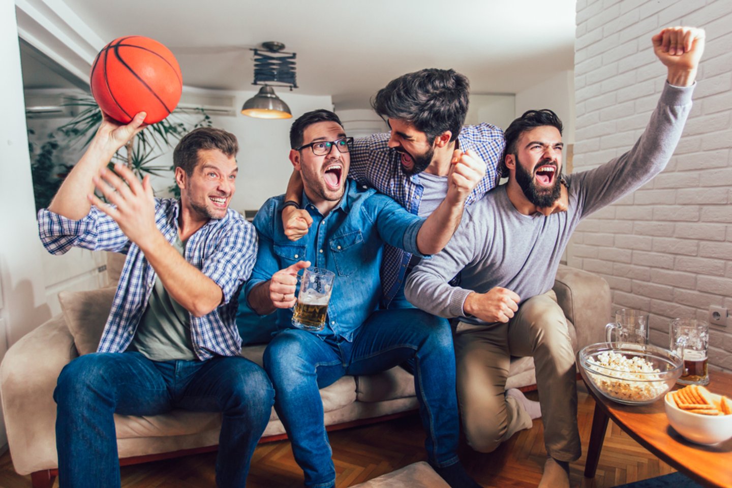 A group of cheering friends on a couch with popcorn, chips and beer in front of them. One is holding a basketball.