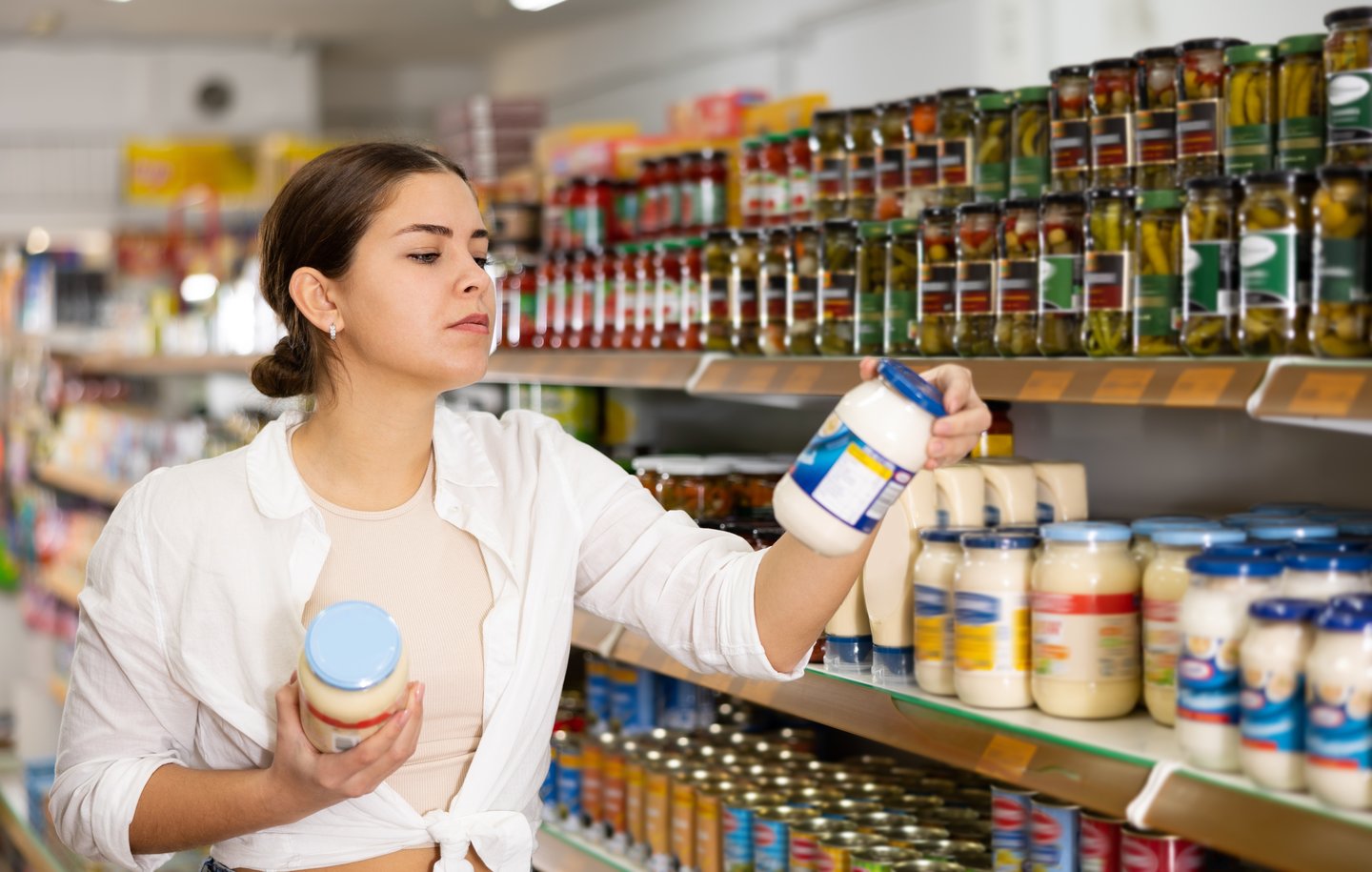 Young dark-haired woman at store shelves holding mayonnaise jar, reading label