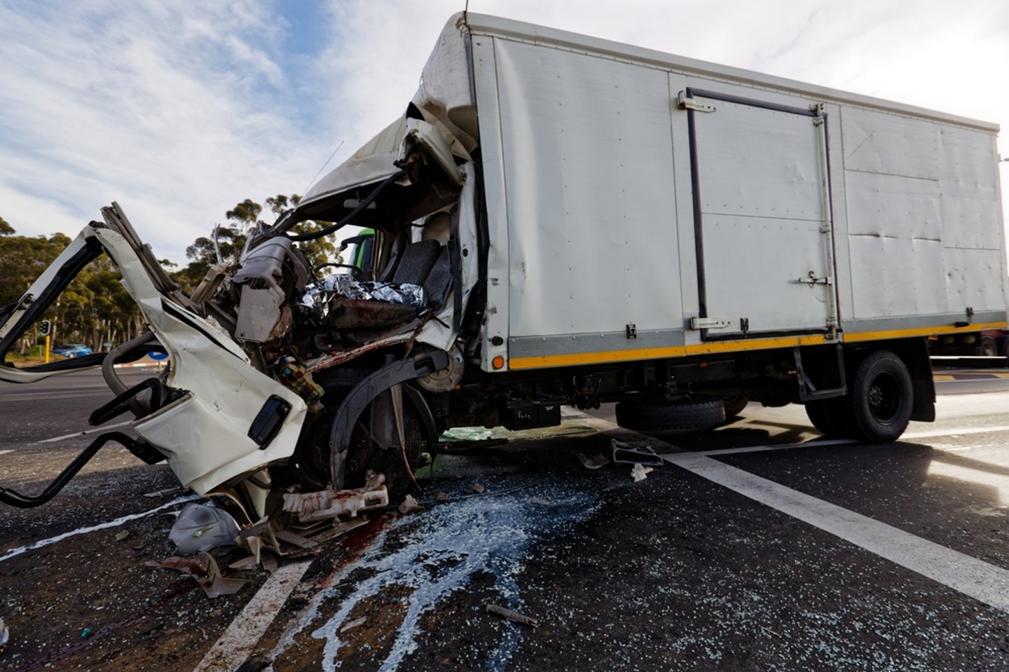 An overtuned truck with its front cab badly damaged in impact; liquid is leaking out from underneath the mangled truck.