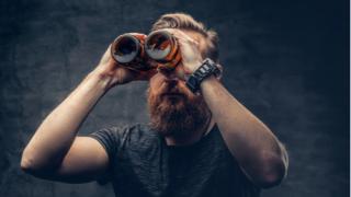 Funny redhead bearded male looking through two craft beer bottle like through binoculars. F