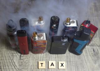 Multiple vaping products with smoke in background. The word "tax" written out in wooden letter box in front of vape products.