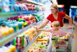 Adult woman pushes a grocery store basket with a child in it, both reach for produce from a grocery store fridge.