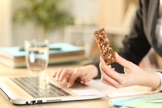 Close up of entrepreneur woman hands holding cereal snack bar working on laptop at home office