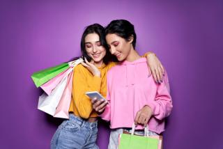 Two happy women friends shoppers holding shopping bags using mobile apps for online shopping standing on purple background. Retail ecommerce fashion sale offers, mall discounts in applications concept