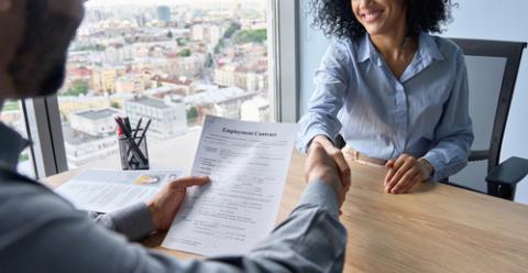Closeup of Indian employer hr holding employment contract job offer hiring welcoming female African newcomer worker manager shaking hands in contemporary office. Getting new job handshake concept.
