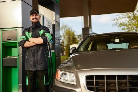Happy bearded man in uniform folding arms and looking at camera with smile, while leaning on pump near modern car on sunny day at gas station