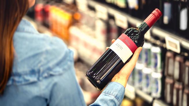 Woman reading the label of red wine bottle in liquor store or alcohol section of supermarket. Shelf full of alcoholic beverages. Female customer holding and choosing a bottle of merlot or sangiovese.