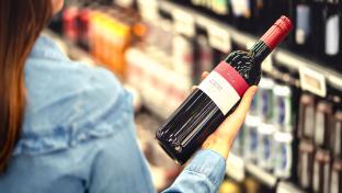 Woman reading the label of red wine bottle in liquor store or alcohol section of supermarket. Shelf full of alcoholic beverages. Female customer holding and choosing a bottle of merlot or sangiovese.