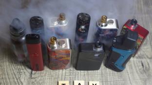 Multiple vaping products with smoke in background. The word "tax" written out in wooden letter box in front of vape products.