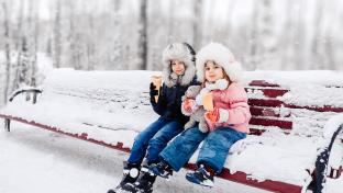 Two young children eat ice cream dressed in winter gear, sitting on a park bench full of snow.