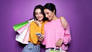 Two happy women friends shoppers holding shopping bags using mobile apps for online shopping standing on purple background. Retail ecommerce fashion sale offers, mall discounts in applications concept